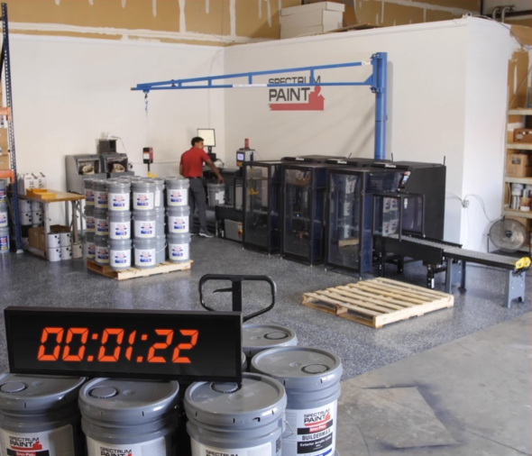 An automated paint mixing line in use at Spectrum Paint, featuring a conveyor system, mixing equipment, and stacked 5-gallon paint buckets, with a visible timer displaying production speed.