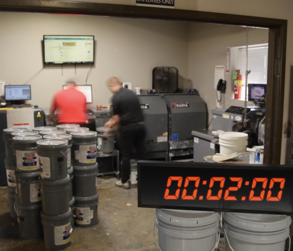 Two workers manually operating Radia paint mixing machines in a production room with multiple stacked paint buckets. A digital timer in the foreground displays '00:02:00'.
