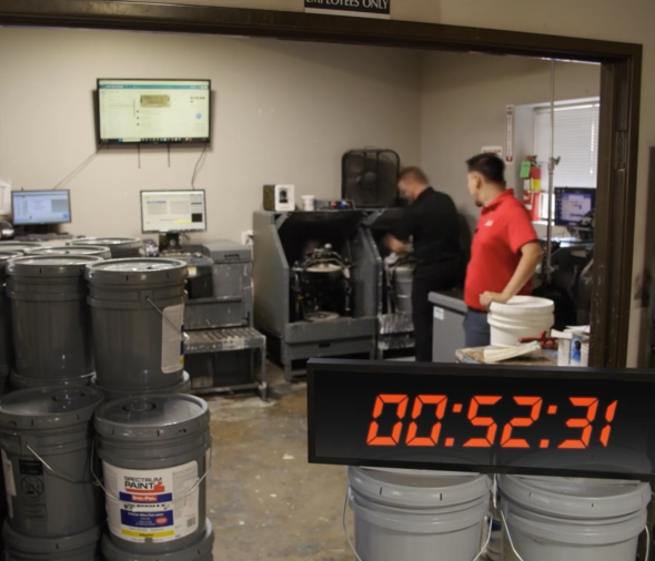 Two employees manually operating paint mixing machines surrounded by buckets, with a digital timer reading 00:52:31 indicating a longer manual mixing process.